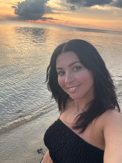 A woman with dark hair smiles at the camera. She is wearing a black strapless top. The location appears to be a beach at sunset in Baan Sri Thanu, Thailand. The water reflects the golden light of the setting sun. The sky contains both orange and grey clouds. Sand is visible at the water's edge.