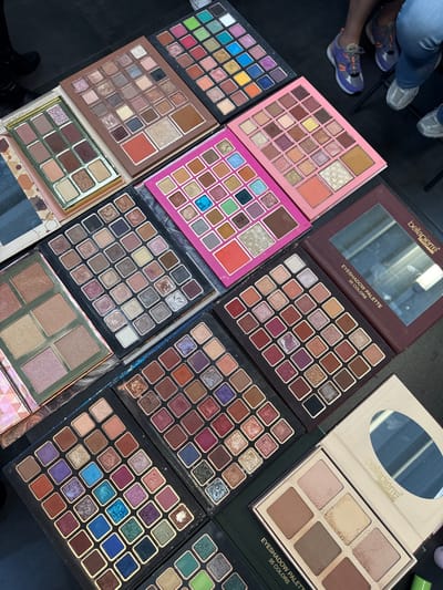 The scene depicts an overhead view of numerous cosmetic eyeshadow palettes laid out on a dark, flat surface in Herentals, Belgium. At least twelve distinct palettes are visible, predominantly featuring multiple square or rectangular pans of various eyeshadows. Color schemes range from extensive neutral and earth tones to vibrant blues, greens, purples, and glitter shades. Brands include 'bellapierre,' visible on one dark brown 35-color eyeshadow palette with a mirror, and another lighter palette containing six larger pans. The upper right quadrant of the image shows the lower legs and feet of at least two individuals; one wears dark blue jeans and purple/black athletic shoes, while another wears light blue jeans and grey athletic shoes. A dark-clad leg is partially visible on the far left.