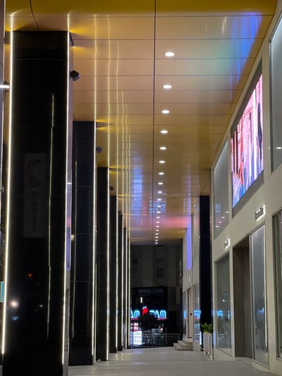 A long, brightly lit corridor or walkway features a series of illuminated steps leading forward. The wall on the left is lined with reflective, gold-toned panels with small, evenly spaced lights. The ceiling above is a grid of panels, some lit with images and others with transparent sections revealing the outside. The steps are black with linear lighting, drawing the eye into the distance where signs and reflections of a building are visible.