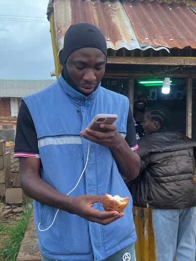 A young Black male wearing a blue vest over a black t-shirt and a black head covering looks down at his phone while holding a piece of food in his right hand. He is standing in front of a small roadside kiosk with a corrugated metal roof. Behind him, a Black man with a beard is smiling and looking at the camera, and a Black woman in a dark puffer jacket and light blue jeans stands next to him. The kiosk appears to be selling electronics, as indicated by a bright green light and several items displayed inside. The background shows some construction materials and a building. The overall scene suggests a casual outdoor setting in a Nigerian city.