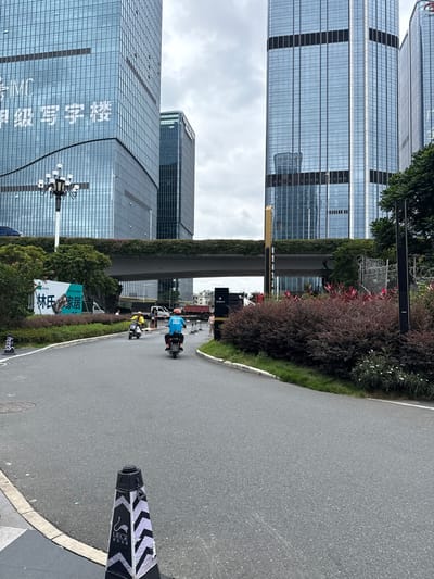 The image depicts a street scene in the Huangpu District, China, dominated by modern high-rise buildings with reflective glass facades. In the foreground, a paved road curves gently to the right, with a black and white striped traffic cone bearing a logo on the left side. Two individuals on scooters are riding on the road, one in a yellow jacket and the other in a blue shirt with an orange helmet. Behind them, a white van and a red truck are visible. Over the road, an elevated walkway or bridge is covered in green foliage, with more buildings visible beyond it. The sky is overcast. The buildings feature numerous windows and vertical metallic elements. Lush green bushes and plants line the sides of the road and the elevated structure. A street lamp with multiple bulbs stands on the left side of the road, near the base of a large building. Signs in Chinese characters are present on a billboard and on some of the buildings.