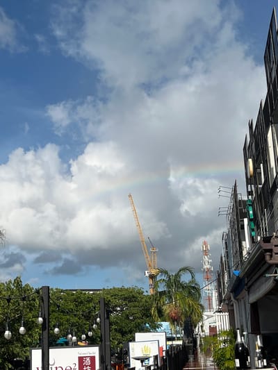 This is a vertical, full-shot photograph of a street scene in Choeng Thale, Thailand, dominated by a bright blue sky with dramatic clouds. A faint rainbow arcs across the upper part of the image. In the midground, a yellow construction crane extends diagonally upwards, and a taller communication tower with red and white sections is visible further back.
The foreground is lined with trees and streetlights, with several signs on poles. One sign in the lower right corner has text in Thai script and a stylized drawing of bananas. Another sign, partially visible, appears to have the word "wine" in English script. The buildings lining the street are a mix of modern structures with dark tiled roofs and balconies, and other commercial buildings with awnings. The scene suggests an urban or commercial area, possibly during or after a rain shower, given the presence of the rainbow and the slightly damp appearance of the ground in some areas. The time of day is likely daytime, with the sun illuminating the clouds and creating the rainbow. There are no people visible in the frame. The overall impression is one of urban development contrasted with natural elements and signs of recent weather.
