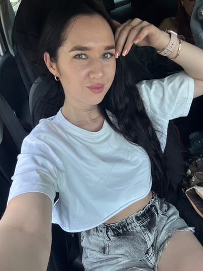 A young woman with dark hair and green eyes is seated in the driver's seat of a car. She is wearing a white crop top and grey denim shorts. The woman is looking at the camera, with her left hand resting on her forehead. She is wearing a ring on her right hand and a smartwatch on her left wrist. The car interior is visible, including the seats and a glimpse of the dashboard. The background suggests an outdoor setting, potentially a parking lot or a street. There is no indication that this photo was taken in Leninsk-Kuznetsky, Russia.