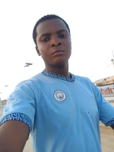 A medium-range, low-angle photograph captures a young man wearing a light blue t-shirt with the Manchester City logo on the left side of his chest. The t-shirt has a patterned blue and white trim around the neck and sleeves. The man has short dark hair and is looking directly at the camera with a neutral expression. 

The background features an urban environment with streetlights and buildings visible. A building with a red and white railing is visible. The location appears to be in Ado, Nigeria. The sky is overcast.