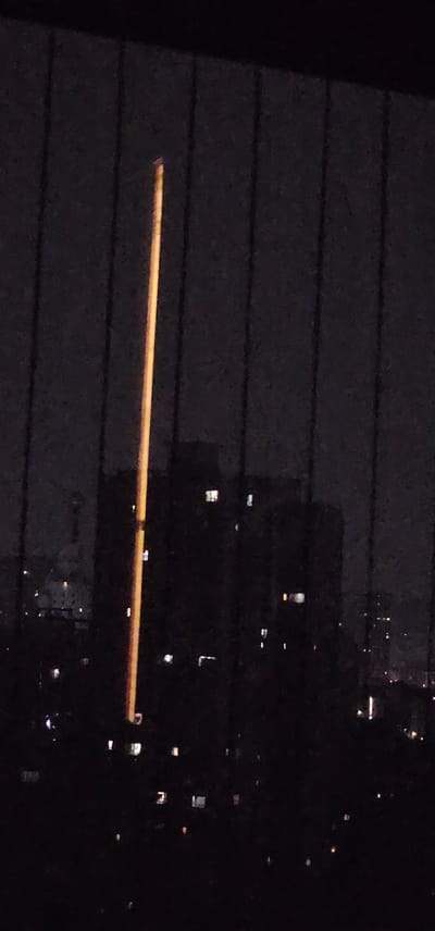 The image shows a view of a city at night, likely from an elevated position such as an apartment window. A tall, slender, illuminated object, possibly a light pole or antenna, dominates the center of the frame and extends vertically from the bottom to near the top. In the background, silhouetted buildings with numerous lit windows are visible, indicating a densely populated urban area. The overall scene is dark, with the light from the central object and the building windows providing the primary illumination. The context of Jiulongpo District, China, suggests this is an urban landscape within that specific geographical location.