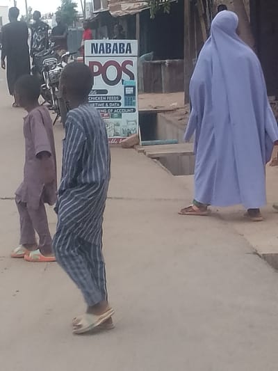 In Pantame, Nigeria, two young boys in traditional attire, one in a purple tunic and trousers, the other in a blue and white striped outfit, walk past a "NABABA POS" advertising sign. Behind them, an adult, cloaked in a light purple garment, walks away from the camera. In the background, several other individuals are visible, along with a motorcycle and market stalls, suggesting a bustling urban environment. The ground is dusty and uneven, typical of a street in Pantame. The overall scene captures a moment of everyday life in this Nigerian city.