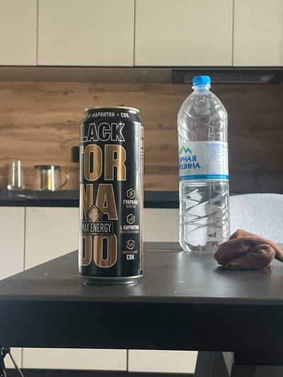 The image shows two beverages on a dark, reflective surface, likely a table or countertop. In the foreground, a black can with gold lettering stands vertically. The can features prominent text that appears to be "BLACK", "OR", and "DO", along with "MAX ENERGY" and various smaller text elements in Russian, including "1-КАРНИТИН", "ГУАРАНА", and "COK". Behind the can, a clear plastic bottle filled with water is lying horizontally. The bottle has a blue cap and a label with blue and green accents, also displaying Russian text that includes "ОРНАЯ" and "РШИНА". A brown cloth is draped over the top of the bottle. The background is blurred, showing vertical white tiles on the left and a wooden texture on the right. The lighting suggests an indoor setting, possibly during the day. There are no people visible in the image.