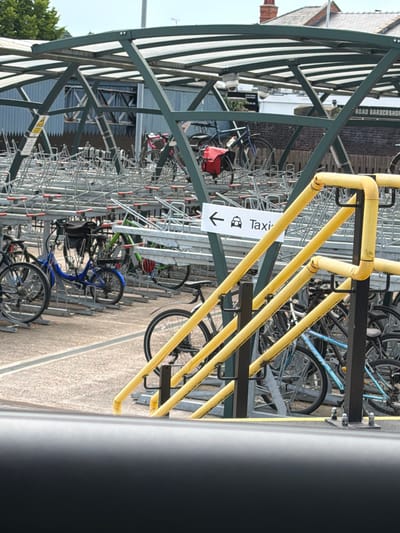 This image depicts an outdoor bicycle parking facility in Cheltenham, United Kingdom. The scene is dominated by a metal structure with yellow tubular beams supporting the roof and providing a framework for bicycle racks. Numerous bicycles are parked, showcasing a variety of styles and colors, including a bright blue bicycle prominently displayed. Some bicycles are equipped with baskets or bags, like the red bag on one of the bikes.

The setting appears to be a sunny day, judging by the bright lighting and clear sky visible in the background. The ground is paved with a light-colored surface. Behind the bicycle racks, glimpses of buildings are visible, including what looks like a brick building with signage. One sign, in white with black text and an arrow, reads "Taxi" and includes an icon of a taxi. Another sign, partially visible, appears to be a yellow warning sign. The overall impression is a functional urban space designed to accommodate cyclists. The composition is vertical, with the structure of the bicycle rack occupying a significant portion of the frame.