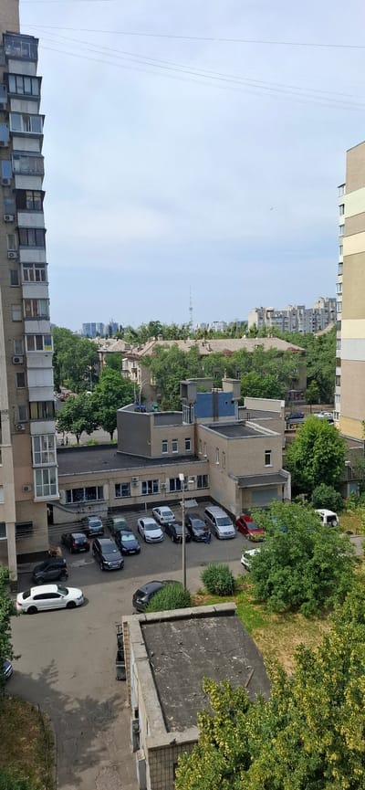 This is an aerial shot of a residential area in Kyiv, Ukraine, featuring a mix of apartment buildings and smaller structures. The foreground is dominated by the side of a tall, multi-story apartment building on the left, characterized by numerous balconies and windows. To the right of this building, a lower, light-colored brick building with a flat roof is visible, with a parking lot in front of it. Several cars are parked in the lot, including a white sedan, a dark-colored sedan, a blue sedan, a silver minivan, and a red car. Further into the background, more apartment buildings and trees fill the landscape, suggesting a densely populated urban environment. The sky is overcast with some faint clouds. A few people are visible on the street and sidewalks. The scene appears to be a typical urban scene during the daytime.