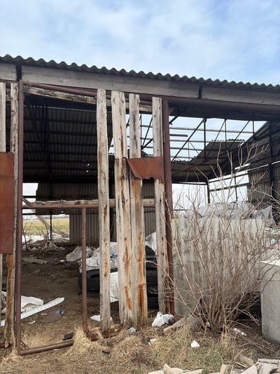 The image shows a dilapidated, open-sided structure located in Bessergenovka, Russia, under an overcast sky. The structure's roof, constructed from corrugated material, is partially intact on the left but exhibits sections of collapse or missing panels on the right, revealing underlying metal framework and a second, lower corrugated roof section. The left side of the structure is partially enclosed by vertical, weathered wooden slats and rusty metal support beams. The interior ground surface is dry earth and sparse dry grass, littered with debris including multiple white industrial bags and at least three black vehicle tires. A leafless, multi-branched bush is visible in the right foreground. Through the structure's open side, a flat, open field with patches of light-colored ground or residual snow extends into the distance under the same cloudy sky. No individuals are present.