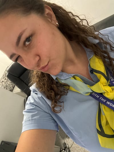 The media file is a close-up, horizontally oriented selfie of a young woman in Aksakovo, Bulgaria. She is wearing a light blue collared shirt, with a yellow and black patterned scarf tied around her neck and extending over her shoulder. A purple lanyard with white text is visible around her neck, partially obscured by the scarf. The woman has long, curly brown hair that falls around her face and shoulders. Her gaze is directed towards the camera, and she appears to be looking slightly to the left. Her expression is neutral or subtly smiling.
The background is blurred but suggests an indoor setting, with hints of furniture and a wall. The lighting is soft, suggesting it might be daytime or an indoor environment with artificial lighting. The overall mood is casual and personal.
Visible text on the lanyard includes fragments of words, possibly "WITH THE WOR". The pattern on the scarf includes yellow and black elements, possibly geometric or abstract.