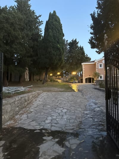 This image depicts an exterior view of a building and its grounds in Boreti, Montenegro. The scene is a stone pathway leading towards a two-story building with a light peach-colored facade. The building has arched doorways and windows with white frames. The pathway appears to be wet, reflecting the lights from the surrounding area. Tall cypress and other trees line the pathway and the grounds. In the background, to the left, a white structure resembling a monument or a tomb is visible amongst the trees. Several lampposts with a warm glow are also present, illuminating parts of the pathway and the greenery. A black wrought-iron gate is partially visible on the right side of the frame, suggesting an entrance to the property. The sky is a clear, twilight blue, indicating that the photo was taken at dusk or dawn. There are no people visible in the image.