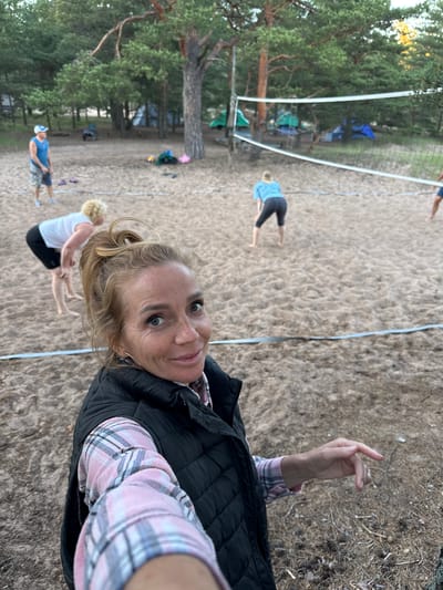 In Ozerki (Krasnodolinskaya), Russia, a group of people are playing volleyball on a sandy court. In the foreground, a woman with blonde hair tied in a ponytail, wearing a pink and white plaid shirt and a black vest, smiles at the camera. She is positioned to the right of the frame, with her left arm extended towards the viewer. In the background, three other individuals are visible. One woman with blonde hair, wearing a white tank top and black shorts, is bent over on the sand. Another woman, also with blonde hair, is in the background, wearing a blue long-sleeved shirt and black leggings, in a volleyball stance. A man wearing a blue tank top and patterned shorts is standing further back on the left side of the court. The court is surrounded by pine trees, and several tents are set up in the background, suggesting a campsite or park setting. A volleyball net stretches across the middle of the court. The ground is covered in sand, with scattered pine needles and cones. The lighting suggests it is late afternoon or early evening.