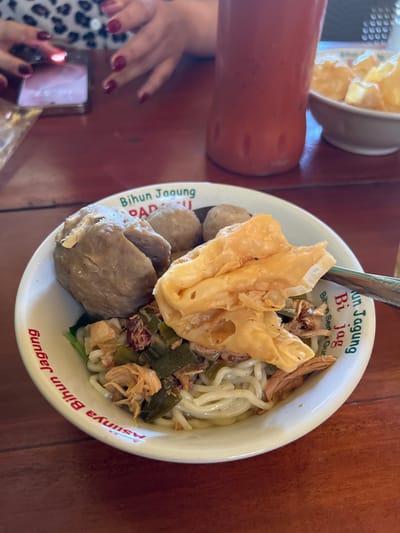 A close-up shot shows a bowl of noodle soup on a wooden table. The bowl has red text printed on its rim. The soup contains meatballs, noodles, and shredded chicken. A fried wonton is perched on top of the noodles. In the background, a person's hands are visible, one holding a smartphone. A bottle of red liquid and a small bowl of snacks are also present. The scene appears to be in Gianyar, Indonesia.
