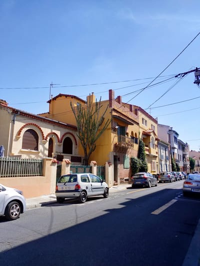 This is a street-level, eye-level, full shot image of a residential street in Perpignan, France, on a clear, sunny day. The perspective is from the sidewalk, looking down the street which curves slightly to the right. The left side of the street is lined with a row of multi-story buildings, primarily residential, painted in shades of yellow, beige, and white, with red tile roofs. The architecture features arched windows, balconies with wrought-iron railings, and some decorative brickwork. A tall, spiky cactus-like plant stands prominently in front of one of the yellow buildings.
The right side of the street is also lined with buildings of similar height and style, also with red tile roofs. A line of parked cars is visible along the curb on the right side of the street. These include a silver Renault Twingo, a grey Volvo, and several other vehicles further down the street, some of which have French license plates. Power lines crisscross overhead, supported by poles. The road surface is asphalt, and a clear shadow is cast across it from the buildings on the left. The sky is a vibrant, cloudless blue. There are no people visible in the frame.