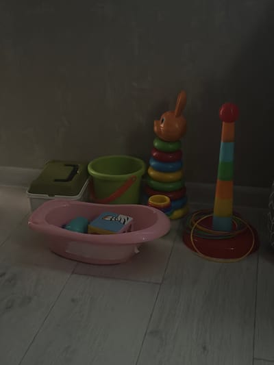 This media file features a collection of children's toys arranged on a light-colored wooden floor. The primary objects are a stacking ring toy with colorful rings and a red base, and a segmented caterpillar toy with an orange head and colorful rings. Next to these, a green bucket with an orange handle and a small green lidded container are visible. A pink bathtub-shaped toy, containing a few smaller colorful objects, sits to the right of the green bucket. The background is a dark, textured wall, possibly a closet or wall surface, which obscures much of the setting. The lighting suggests an indoor environment, likely a room with low ambient light, possibly an evening or a dimly lit area. The overall scene depicts a play area, hinting at the presence of children. No people are visible in the image. There is no visible text in the media file. The location context is Krasnoyarsk, Russia.
