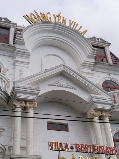 The image shows the upper facade of a white building with gold lettering that reads "HOÀNG YẾN VILLA". The building features ornate architectural details, including columns, arched windows with red frames, and decorative molding. Below the "HOÀNG YẾN VILLA" sign, another red and gold sign reads "VILLA - RESTAURANT". The sky above is overcast. The image appears to be taken from a street-level perspective in Lâm Viên - Đà Lạt, Vietnam. There are no people or animals visible in the image.
