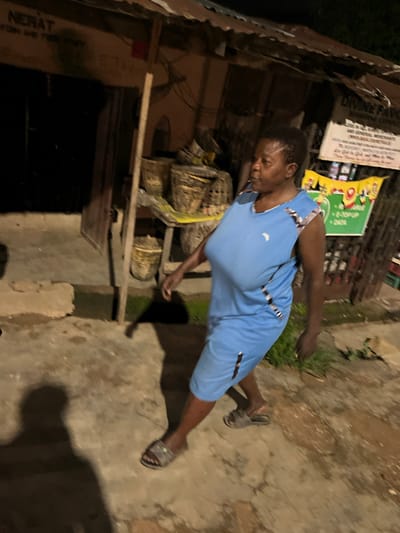 A woman is walking in front of a shop in the city of Gura Top, Nigeria. The woman is wearing a blue dress and sandals. The shop is made of corrugated iron and wood, and it has a sign that says "NERAT FISH AND FOOD MART". There are baskets of produce outside the shop. The ground is made of concrete, and there is a shadow of the woman on the ground. The lighting is dim, suggesting it is either nighttime or dusk.