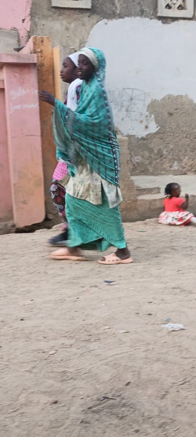 This outdoor image captures a daytime street scene in Rangaza, Nigeria. In the foreground, two young women or girls are walking from left to right on a sandy path. The person in front wears a light teal head covering and a matching long skirt over a lighter top, paired with light peach open-toed sandals. Her dark-skinned face is visible in profile, showing a slight smile. Partially obscured behind her is another dark-skinned individual wearing a white head covering, a light top with a pink pattern, and dark shoes.
On the right side of the mid-ground, a small dark-skinned child sits on the sandy ground or a low step. The child is dressed in a bright red top and a white skirt with a red floral pattern, and has small braids or pigtails. They appear to be looking downwards.
The background consists of weathered walls and structures, suggesting a residential or village setting. On the far left, a light pink column or pillar shows faint, handwritten text, which is mostly illegible but includes fragments resembling "Gulans" and "Bee." The ground is sandy or dirt, with minor debris scattered throughout. The lighting indicates it is daytime, with diffused light and no harsh shadows.