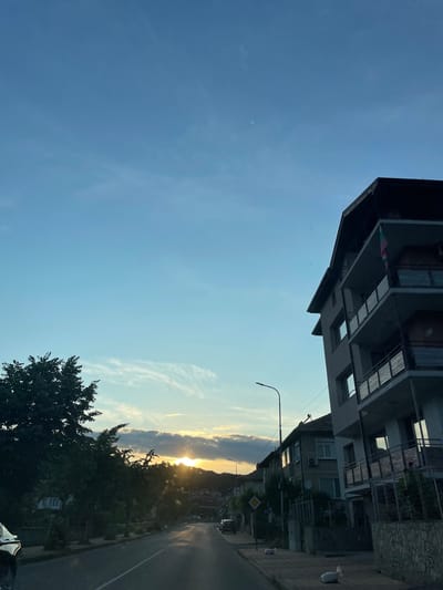 This is a vertical, medium shot of a street scene in Zlatograd, Bulgaria, during what appears to be sunset or sunrise. The sky is a clear blue with wispy clouds, and the sun is visible peeking through a large cloud formation, casting golden rays. On the left side of the frame, a modern building with dark grey siding and multiple balconies is prominently featured, partially obscuring the sky. On the right side, a paved road stretches into the distance, lined with trees and more buildings. A street lamp is visible on the right, and a small, diamond-shaped yellow road sign is also present. The overall mood is serene, with the warm glow of the sun contrasting with the shadows cast by the buildings and trees. No people are clearly visible in the image.
