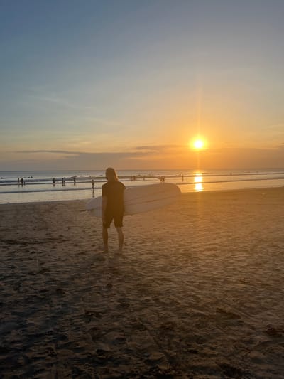 A person stands on a sandy beach, holding a surfboard. They are facing the ocean, with the sun setting in the background. The sky is a gradient of orange and blue, and the ocean is calm with small waves. Several other people are visible in the water and on the beach. The scene appears to be a typical sunset at Kuta Beach, Bali, Indonesia, a popular spot for surfing. The sand is a warm, golden color, reflecting the light of the setting sun. The overall atmosphere is serene and peaceful.