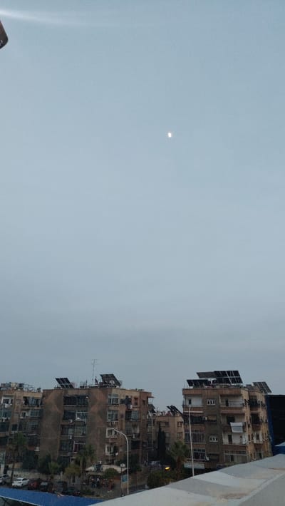 This image depicts an urban landscape within Barza Municipality, Syria, captured from an elevated perspective during twilight or overcast conditions. The upper portion of the frame features a pale grey sky with a distinct, slender crescent moon visible in the upper-middle. The lower two-thirds of the image are dominated by multiple multi-story residential buildings constructed from light brown or tan materials.

These buildings exhibit numerous balconies, some with visible items such as laundry or storage, and a pattern of windows, some dark, others reflecting ambient light. A prominent feature across the rooftops of most visible structures is the presence of multiple dark, rectangular solar panel arrays, alongside various satellite dishes and antennae. Exterior air conditioning units are mounted on several building facades.

Between the buildings, a street or thoroughfare is visible, populated with parked vehicles, including several light and dark-colored cars. Ground-level sections of the buildings show indications of commercial activity, such as small storefronts or shop entrances. Scattered urban vegetation, including palm trees, is interspersed among the structures. A street light pole with a luminaire extends upwards from the left midground. The immediate foreground shows the top edge of a light-colored, textured barrier or rooftop from which the photograph was taken. No clearly identifiable individuals or specific actions are discernible.