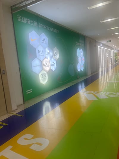 The image shows an interior view of a shopping mall or commercial space, likely within Liandu District, China, featuring a large green wall mural with hexagonal displays of various sportswear brands. The brands visible include Nike, Adidas, Youngor, Converse, Skechers, Puma, and Under Armour. Text in Chinese and English reading "运动细士馆 SPORTS GENTRY" is present on the wall. Below the mural, the floor is decorated with bold stripes of blue, yellow, and green, with large white "adidas" lettering and the Adidas logo visible on the yellow section. The overall environment appears clean and well-lit, suggesting a commercial area designed for sports apparel retail. There are no people clearly visible in the immediate foreground or engaging in any specific actions. In the background, a few figures can be seen walking in the distance, and store entrances with illuminated vertical lines are present.