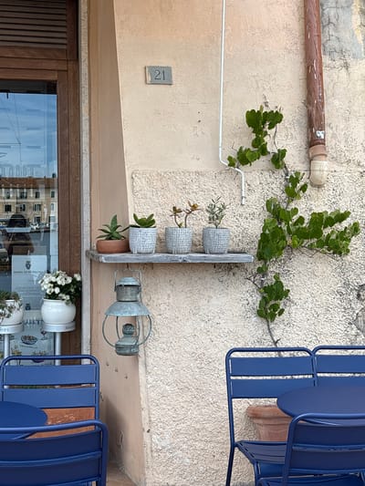 This image captures a quaint street view, likely an exterior wall of a building in Portoferraio, Italy. The scene features a textured, light beige stucco wall adorned with various elements.

Running horizontally across the upper part of the wall is a weathered brown pipe, with a small white conduit visible below it, extending diagonally. A vibrant green vine with broad leaves grows along the wall, guided by a thin wire.

Mid-frame, a rustic wooden plank holds three small grey ceramic pots, each containing a different succulent or herb plant. Below these, an old-fashioned, galvanized metal lantern hangs, adding a touch of vintage charm.

On the left, a grey rectangular plaque displays the house number "21." The lower section of the image reveals a dark wooden door or window frame, with a large glass pane reflecting the surrounding environment. In the reflection, parts of other buildings, possibly the sky, and interior details are visible. Several signs or menus with Italian text are discernible in the reflection, including "BADIA DI MORRONA" (a Tuscan winery), "PANZANELLA" (a Tuscan bread salad), "DI PESCE" (of fish), and "BANFI" (another Italian winery). A white pot filled with small white flowers sits on a ledge in front of the glass.

To the right, two bright blue metal outdoor chairs, typical of cafe or bistro seating, are stacked, with parts of their slatted backs and seats visible. A terra cotta pot is partially seen beneath the chairs.

The bright lighting suggests it is daytime, and the overall impression is peaceful and inviting, characteristic of a Mediterranean setting. There are no people visible, implying a quiet moment.