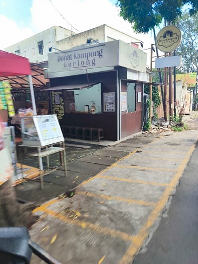 A street-level, slightly blurry view of a small, brown wooden food stall in Bandung, Indonesia (latitude: -6.868, longitude: 107.581). The stall is labeled "Donat Kampung Gerlong" and features a white sign with a picture of a donut on it. The stall has a window and a small counter. There are four metal stools in front of the counter. A metal stand with a glass case and a white sign sits in front of the stall, facing the street. To the right of the stall is a sidewalk with yellow painted lines, and a brown wooden pole with a circular sign that reads "Donat Kampung Gerlong" on it. To the left of the stall is a red tent and a building. The scene is taken from the sidewalk and there are no people in the picture. There is a small, leafy tree to the right of the stall. The ground is paved with concrete. The sky is bright, indicating it is daytime. The image appears to be taken from a moving vehicle.