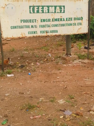 The image shows a roadside construction sign indicating a project undertaken by the Federal Roads Maintenance Agency (FERMA). The project is identified as "Engr. Emeka Eze Road," with Fractal Construction Co. Ltd. listed as the contractor. FERMA Abuja is noted as the client. The sign is mounted on a metal pole. In the foreground, the ground is covered with dirt, litter, and some sparse vegetation. A chicken is visible near the left edge of the image.
