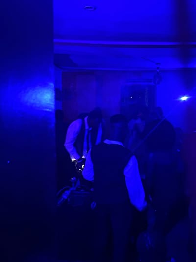 In the city of Durumi, Nigeria, a group of people are gathered in a dimly lit indoor space, illuminated by intense blue lighting. Several individuals, appearing to be young men dressed in formal attire, are visible. One person in the foreground is wearing a white shirt and a dark vest, with a dark tie. Another individual, also in a vest and white shirt, is partially obscured but seems to be bending over, possibly interacting with equipment on the floor. The environment appears to be a social gathering or event, possibly a party or celebration, with a ceiling fan visible in the upper portion of the frame. The blue lighting creates a dramatic and somewhat hazy atmosphere, with some light beams or reflections visible. The overall impression is that of a lively, perhaps festive, indoor scene.