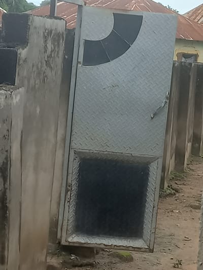 The image shows a weathered, diamond-patterned metal door set into a rough, concrete structure. The door has a square glass window on the right side, also framed with the same diamond-patterned metal. On the left side of the door, there is a quarter-circle shaped section that is black, also with a diamond-pattern. The door appears to be part of an outdoor, possibly dilapidated, building or enclosure. The surrounding ground is dirt and dry vegetation. In the background, part of a red corrugated metal roof is visible, along with more of the concrete structure and some green foliage. The lighting suggests it is daytime, with no strong shadows indicating a specific time. The setting appears to be a somewhat neglected urban or semi-urban area in Mararaba, Nigeria. There is no visible text in the image.