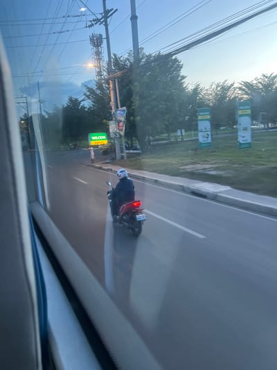 The image shows a person riding a motorcycle on a road. The rider is wearing a helmet and dark clothing, and the motorcycle has a license plate that is partially visible. In the background, there are trees and utility poles. A sign with green and yellow lettering is also visible. The sky is overcast, suggesting it might be a cloudy day or near dusk. The scene appears to be in an urban or suburban area, possibly along a main road given the presence of a median and multiple lanes. The overall impression is that of a casual commute or travel on a motorcycle. The sign reads "WILCON DEPOT".
