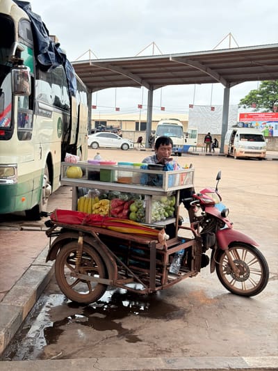 A man is sitting behind a mobile cart filled with fruits and drinks. The cart is attached to a red motorcycle. The scene is outdoors, under a covered walkway. Several buses and vans are parked in the background. The ground is wet and appears to be a bus station or transit area. The man is looking down, possibly at his products or a customer. There are no direct interactions visible between the man and other people. The environment suggests a busy transportation hub.