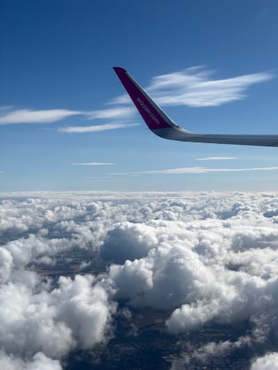 An airplane wing, branded with "WIZZ AIR," extends into a clear blue sky dotted with wispy white clouds. Below the wing, a vast expanse of puffy white clouds stretches to the horizon, partially obscuring the patchwork of land and buildings visible beneath. The scene captures the perspective from an airplane window, offering a view of the earth from above. The specific location in Rakovica Urban Municipality, Serbia, cannot be determined from the image provided.