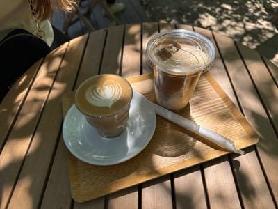 A wooden table with slats stands outdoors. Sunlight dappled with tree shadows illuminates the table. On the table sits a wooden tray with a latte in a glass cup on a white saucer, an iced coffee in a plastic cup with a lid, and a white straw. A person partially visible in the upper left corner has a black handbag with a gold chain. The location is Dongcheng District, China.