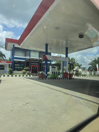 The image captures a view of a Pertamina gas station located in Seumadam, Indonesia. The gas station has several pumps advertising Pertamax Turbo, Pertamax 92, and Dexlite fuel options.  A building nearby is labeled “PT. Putra Tamiang SPBU 14.244.475 Seumadam". A POS Security booth is partially visible. The photograph appears to be taken from inside a vehicle.