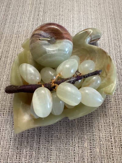 A decorative stone sculpture of an apple and grapes rests on a textured surface. The apple, with swirls of red and green, is positioned at the top, appearing to be attached to a green, leaf-shaped dish. The dish is filled with several smooth, oval-shaped stone grapes, some of which are clustered around a dark, fibrous stem. The background is a woven fabric with a neutral, textured pattern. The scene is lit from above, creating subtle shadows. The image does not contain any information about the city of Tashkent, Uzbekistan.