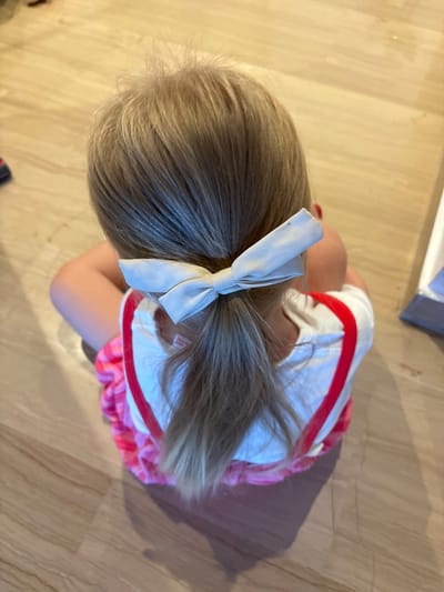 A young child with blonde hair tied into a ponytail with a light-colored bow sits on a wooden floor. The child is wearing a white shirt with red trim and a pink and white patterned skirt. The scene appears to be indoors, possibly in a home. There is no discernible information within the image to determine its location in the city of València, Spain.