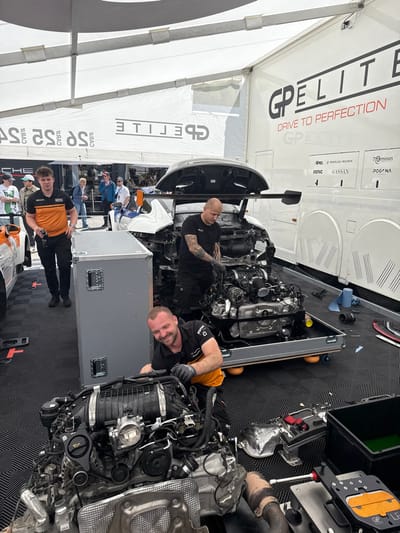 The image shows a scene from a racecar pitstop area located in Stavelot, Belgium, near Circuit de Spa-Francorchamps. Three men are working on a disassembled racecar, specifically the engine. The engine is out of the car and resting on a toolbox. A white racecar with its hood raised can be seen in the background, along with a large white trailer with a sign for "GPELITE, DRIVE TO PERFECTION." The men are wearing racing gear, including fire suits and gloves. The two men working on the engine are kneeling while the third man is standing nearby. The ground is covered in a black patterned mat.