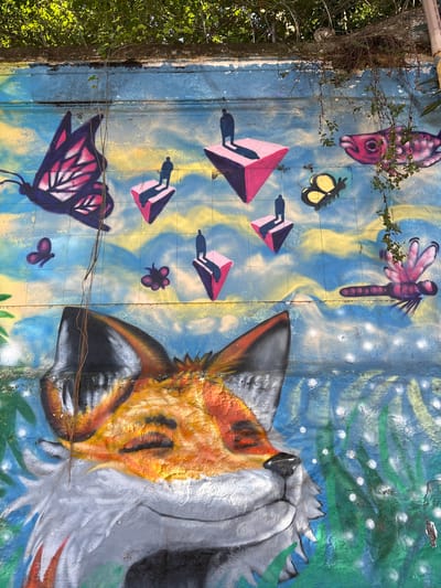 This is a photograph of a colorful mural painted on a textured, tiled wall. The mural features a variety of stylized creatures and geometric shapes. On the right side of the frame, a large, partially depicted animal, likely a fox or wolf, with orange, black, and white fur is the dominant subject. Its head is turned away from the viewer, with its snout and fluffy mane visible. To the left of the animal, the mural depicts several abstract, kite-like shapes in pink and purple, with dark shadows suggesting movement. Other elements include a pink dragonfly, a yellow butterfly, a pink fish with its mouth open, and smaller purple butterfly-like shapes and flowers. The background is a vibrant blend of blues and yellows, with green and white accents that could represent foliage or abstract patterns. The wall itself appears to be outdoors, possibly a boundary or exterior wall, as indicated by the presence of greenery and vines growing from the left edge of the frame. The lighting suggests it is daytime, with shadows from the vines falling on the mural. There are some wires strung across the lower portion of the image, obscuring parts of the mural. The mural has a whimsical and fantastical theme, with a bright and energetic color palette. No text is visible in the image. The context provided indicates the location is São Paulo, Brazil.