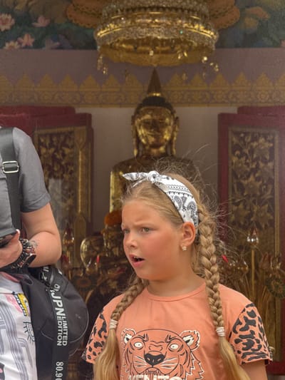 A young girl with blonde hair, styled in two braids and adorned with a black and white bandana, stands in the foreground. She wears an orange t-shirt with a tiger graphic and leopard print sleeves. Her mouth is open, and she appears to be speaking or reacting to something. Beside her, partially visible on the left, is a person wearing a grey t-shirt and a black bag. Behind them, a golden Buddha statue is visible within a temple-like structure, decorated with ornate gold and purple accents. The background also features intricate gold carvings on red panels and a golden canopy with dangling elements. This scene is located in Chalong, Thailand.