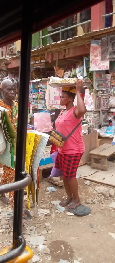 In the Fegge market, Nigeria, a woman in a pink shirt and red plaid skirt balances a tray of goods on her head. She has a patterned shoulder bag and wears flip-flops. A man in a patterned shirt stands next to her. The area is littered, and shops with various items line the street.