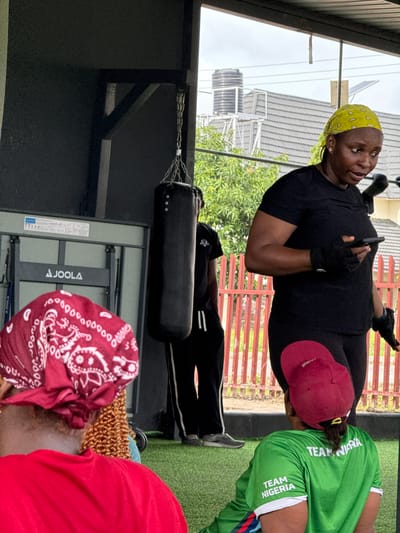 In Iddo Tudunwada, Nigeria, an outdoor gym area features a punching bag hanging from a metal support. A woman in a black t-shirt and leggings, wearing a yellow headscarf and fingerless gloves, stands holding a smartphone. To her left, a person in black athletic wear with white stripes on the pants stands near a table tennis table. In the foreground, the back of a person wearing a red t-shirt and a red bandana is visible, and to their right, another person is seated wearing a green "TEAM NIGERIA" shirt and a maroon baseball cap. The ground is covered with green artificial turf, and a red metal fence is visible in the background.
