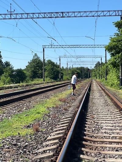The image shows a person standing on railroad tracks in a rural setting, likely in Ureki, Georgia. The person, wearing a white t-shirt and dark shorts with red trim, is captured mid-motion, suggesting they are engaged in an activity such as dancing or posing for a photograph. The tracks stretch into the distance, flanked by lush green trees on one side and a network of overhead power lines on the other. The sky is clear and blue, indicating a sunny day. The overall scene appears to be peaceful and somewhat isolated. There is a red traffic light visible in the distance, positioned over the tracks, and a white sign with the number "2" on it near the person. The time of day appears to be daytime, with bright sunlight casting shadows. The image is oriented vertically, with the person and tracks appearing to run from the bottom right to the top left of the frame.
