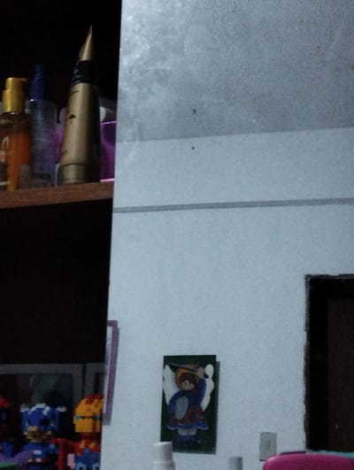 The image depicts an indoor scene, likely a bedroom or living space, in Santa Ana, Venezuela. On the left side of the frame, a dark wooden shelf displays several items. Among these are three bottles: one with an orange liquid and a gold cap, another with a purple liquid and a blue cap, and a third item that appears to be a gold-colored cosmetic tube or bottle with a black band. To the right of the shelf, a light-colored wall dominates the view. A horizontal gray line is visible on this wall, roughly one-third of the way down from the top. Lower on the wall, a framed picture of a child-like figure, possibly an angel, is displayed. The figure has brown hair, a blue tunic, and a red skirt, and is holding a shield and sword. Below the framed picture, two small, light-colored electrical outlets are visible on the wall. In the bottom right corner of the image, a dark doorway is partially visible, suggesting an adjacent room. The overall lighting is dim, with the shelf area being particularly dark. The room appears to be tidy, with no obvious signs of disarray or immediate activity.