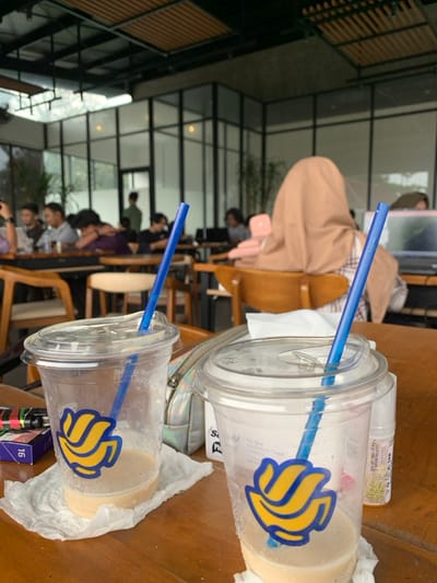 The image shows two plastic cups of iced coffee with blue straws, on a wooden table, at a cafe in Malang, Indonesia, located at -7.966, 112.62. One cup is almost empty, while the other is nearly full. The coffee cups have a sticker on them, featuring a blue and yellow logo, and a white lid with a clear plastic dome. A white paper napkin is under each cup. A woman wearing a light brown hijab is sitting in the background. There is a small pack of cigarettes with a white box and black writing on the table next to the cups. The cafe appears to have a modern design, with a high ceiling and large windows. The cafe is nearly empty. The image focuses on the two iced coffee cups and the woman sitting in the background, with other details out of focus.