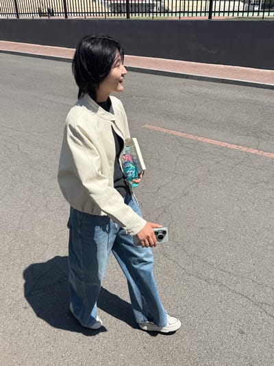 This outdoor shot, taken in Zhongtun, China, features a young person standing on a paved surface under bright sunlight. The individual has short, dark hair and is dressed in a cream-colored or off-white jacket, a dark top, blue jeans, and white sneakers.

They are holding a light-colored smartphone with a visible camera array in their right hand. In their left arm, they cradle a clear plastic bottle containing a pinkish liquid. The bottle's blue label prominently displays the English word "ICE" and Chinese characters which read "冰糖雪梨" (Bīngtáng xuělí), translating to "Rock Sugar Pear." A small, white rectangular object, possibly a book or container, is also held in their left arm.

The setting is an asphalt surface, likely a pathway or parking lot, characterized by its gray texture and a faded orange-red painted line. A light brown brick curb runs alongside, separating the main paved area from a darker, possibly paved, section adjacent to a black metal fence. Strong, clear shadows are cast on the ground, indicating a bright, sunny day. The person is looking upwards or to the side, with a neutral and relaxed expression.
