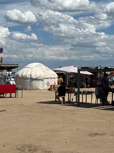 The image depicts an outdoor market scene in Бурсоль, Russia. In the foreground, a sandy ground stretches across the frame, with scattered debris and footprints. To the left, a red market stall with white bags is partially visible, with a large white yurt situated behind it. The yurt is adorned with red decorative patterns. Further to the right, several market stalls are set up under makeshift roofs, some with umbrellas providing shade. People are seen interacting within these stalls, some sitting on stools, while others appear to be engaged in transactions or observing the market. In the background, vehicles and more market structures can be observed. The sky above is filled with dramatic clouds, suggesting a dynamic weather day. A Russian flag is visible on a flagpole to the far left, confirming the location in Russia.