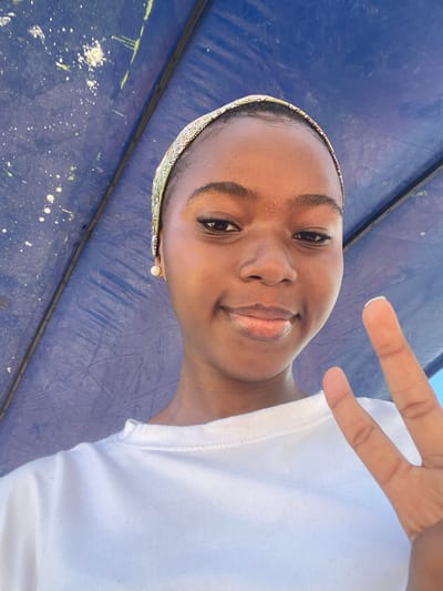 A young Black woman is seen in a close-up selfie. She wears a white t-shirt and a patterned headband. She is making a "peace" or "victory" sign with her fingers. She also wears a pearl earring. The background appears to be a blue tarp or surface with some splattered paint spots. It is not possible to determine the location in Dar es-Salaam, Tanzania from the provided image.